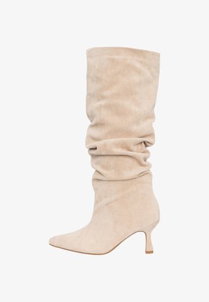 Bota holgada de ante beige hasta la mitad de la pantorrilla, con punta puntiaguda y tacón kitten corto, vista de perfil sobre un fondo blanco.