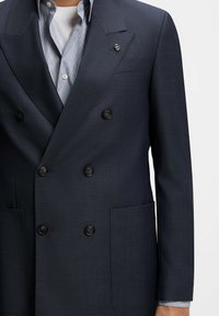 Uomo che indossa un blazer doppiopetto blu scuro con bottoni neri sopra una camicia a righe azzurra e una maglietta bianca sottostante, volto non visibile.