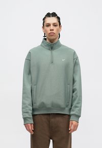 Šviesiai žalia zip-up džemperis su aukštu apykakle, su mažu baltu Nike logotipu ant krūtinės ir dviem šoninėmis kišenėmis.