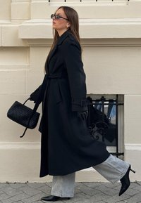 Svart lång kappa med bälte, kombinerad med svarta handskar, eleganta solglasögon och en flätad svart handväska. Ljusblå utsvängda jeans och spetsiga klackar.