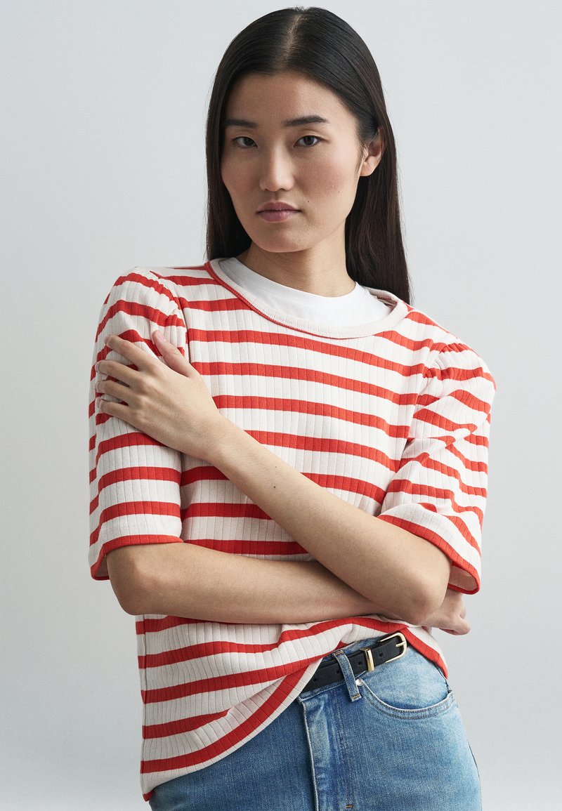 Κόκκινη και λευκή ριγέ κοντομάνικη πλεκτή μπλούζα με ραβδωτή υφή, συνδυασμένη με μπλε τζιν και μαύρη ζώνη.