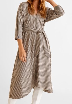 Kobieta w beżowo-brązowej sukience midi w kratę z rękawami do łokci i paskiem do wiązania, w białych botkach.