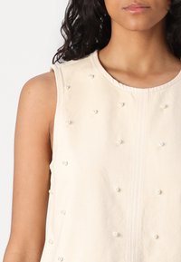 Top senza maniche beige con una finitura testurizzata, decorato con perle distribuite uniformemente sulla parte anteriore e una scollatura rotonda.