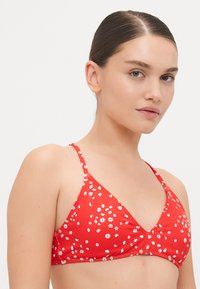 Rød bikinitop med trekantet design og tynne stropper, prydet med hvite blomster mønstre og en glatt, skinnende tekstur.