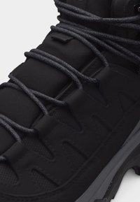 Nahaufnahme eines schwarzen Wanderschuhs mit schwarzen, gemusterten Schnürsenkeln, die durch zickzackförmige Ösen auf strukturiertem Stoff gezogen sind.