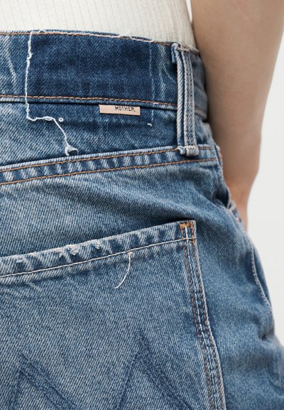 Οπίσθια όψη ατόμου που φοράει μπλε τζιν με την ετικέτα της μάρκας "Mother" κοντά στη μέση και ένα λευκό ribbed τοπ που έχει μπει μέσα.