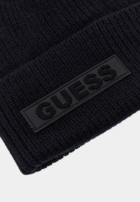 Berretto a coste nero in maglia con una toppa rettangolare in gomma nera che mostra il logo "GUESS" in lettere in rilievo. Design semplice e testurizzato.