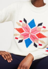 Gestrickter cremefarbener Pullover mit einem farbenfrohen geometrischen Sternmuster in Pink, Blau, Rot, Schwarz und Beige sowie gerippten Bündchen und Kragen.