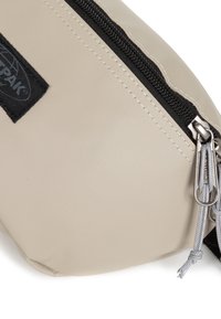 Pochette beige con zip realizzata in materiale liscio e resistente. Presenta accessori neri, una patch a logo rettangolare e dettagli con zip grigia.