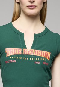 Zelený žebrovaný top s krátkým rukávem, s výstřihem do V a červeno-žlutým grafickým textem: "TRUE RELIGION FASHION FOR THE SENSES SECTION ROW SEAT."