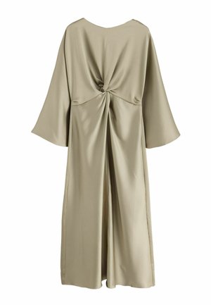 Femme en robe fluide olive à manches larges tient une pochette beige et porte des talons hauts métalliques dorés sur fond vert foncé.