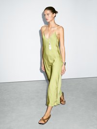 Olive green, sleeveless jumpsuit in lightweight fabric with a V-neck, falling mid-calf. Paired with strappy brown sandals and layered silver jewelry.