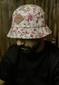 Hatt med blomstermønster i off-white med rosa blomster, med en brun diamantmerke. Mykt stoff med buet skygge.