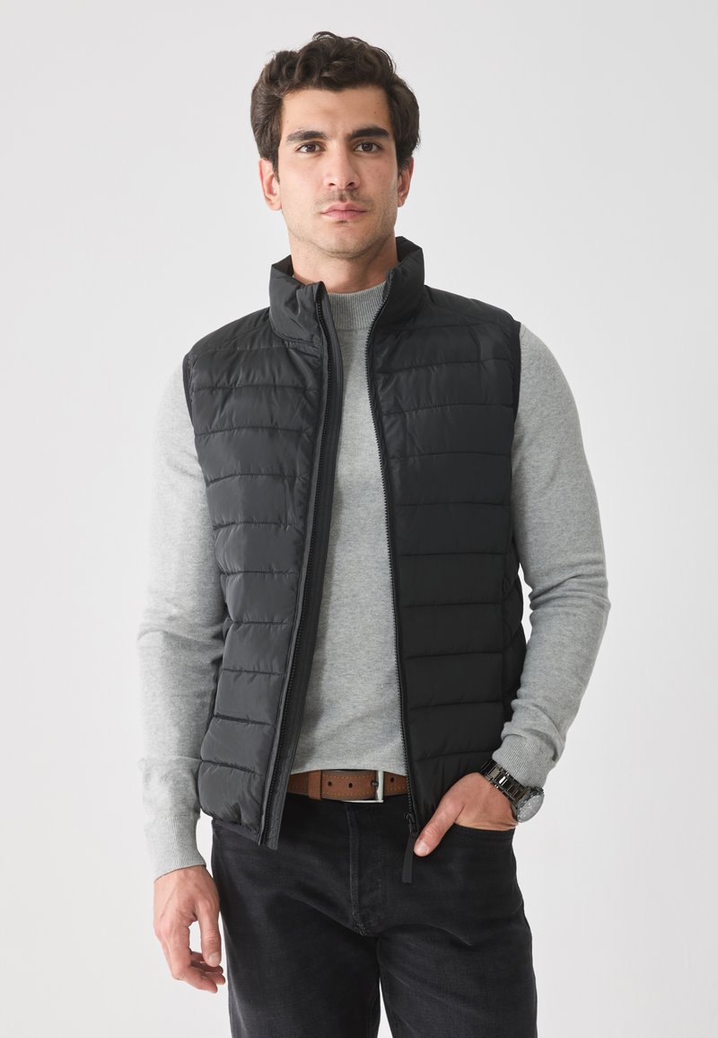 Chaleco acolchado negro con un cuello alto y un patrón de costura horizontal, usado sobre un suéter gris claro, combinado con jeans oscuros y un reloj.