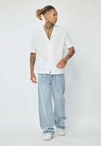 Chemise blanche à manches courtes avec une texture subtile, associée à un jean large délavé bleu clair, présentant des déchirures verticales et des coutures blanches.