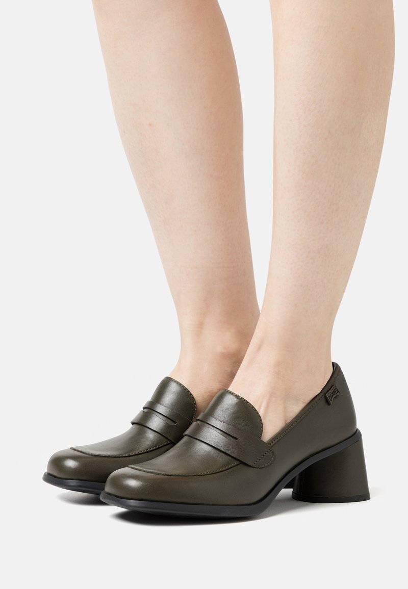 Mocassins en cuir vert olive foncé à talon bloc portés aux pieds, avec une bride sur le dessus et un bout arrondi.