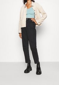 Persona che indossa una giacca in corduroy beige chiaro, un top corto blu chiaro, jeans neri a vita alta e stivali neri spessi alla caviglia, in piedi con una mano sulla vita.