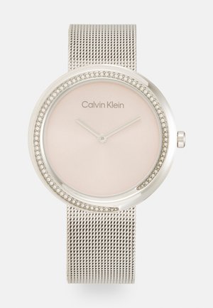 Montre avec un bracelet en maille argentée, un cadran rond rose et une lunette ornée de petites accents métalliques. Présente des aiguilles d'heure et de minute minimalistes.