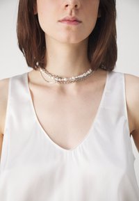 Collana a tre strati composta da catene d'argento con perle bianche di diverse dimensioni e perline allungate, indossata su un top di raso bianco.