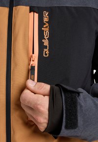 Hand, die den Reißverschluss eines braunen und schwarzen Jackets mit orangefarbener "Quiksilver"-Logo und grauem Ärmel mit schwarzem Klettverschluss anpasst.
