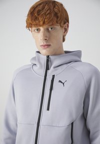 Felpa zip con cappuccio grigio chiaro, colletto alto, dettagli neri e un piccolo logo nero, realizzata in tessuto liscio con un design casual.
