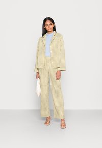 Conjunto de fato em padrão xadrez amarelo e verde, com um casaco ajustado e calças de perna larga, combinado com uma blusa azul clara e sandálias brancas com tiras.