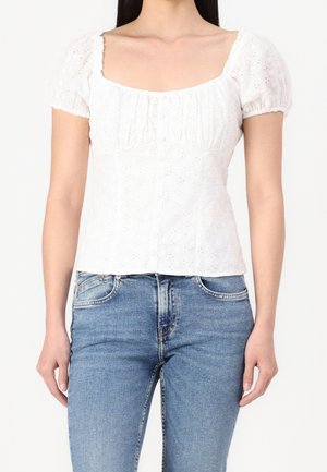 Vrouw draagt een witte korte mouwen blouse met opengewerkt patroon en pofmouwen en blauwe high waist jeans tegen een effen achtergrond.