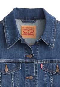 Chaqueta de mezclilla en azul oscuro con un collar clásico, dos bolsillos en el pecho, botones de cobre y una etiqueta de marca. La tela presenta una ligera textura y costuras.