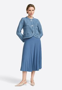 Giacca blu trapuntata con scollatura rotonda e chiusura con bottoni, abbinata a una gonna blu plissettata e fluttuante e scarpe slingback di colore chiaro.