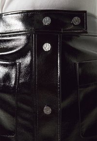 Jupe en cuir verni noir présentant une taille haute, une texture lisse, des poches avant et des boutons-pression métalliques avec un logo.