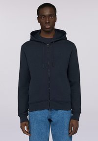 Námořnická mikina na zip z texturované tkaniny, s kapucí na stažení, dlouhými rukávy a decentním logem na hrudi. V kombinaci s modrými džíny.