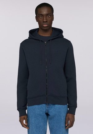 Marineblauer Reißverschluss-Hoodie aus strukturiertem Stoff, mit Kapuze mit Zugband, langen Ärmeln und einem dezenten Logo auf der Brust. Kombiniert mit blauen Jeans.