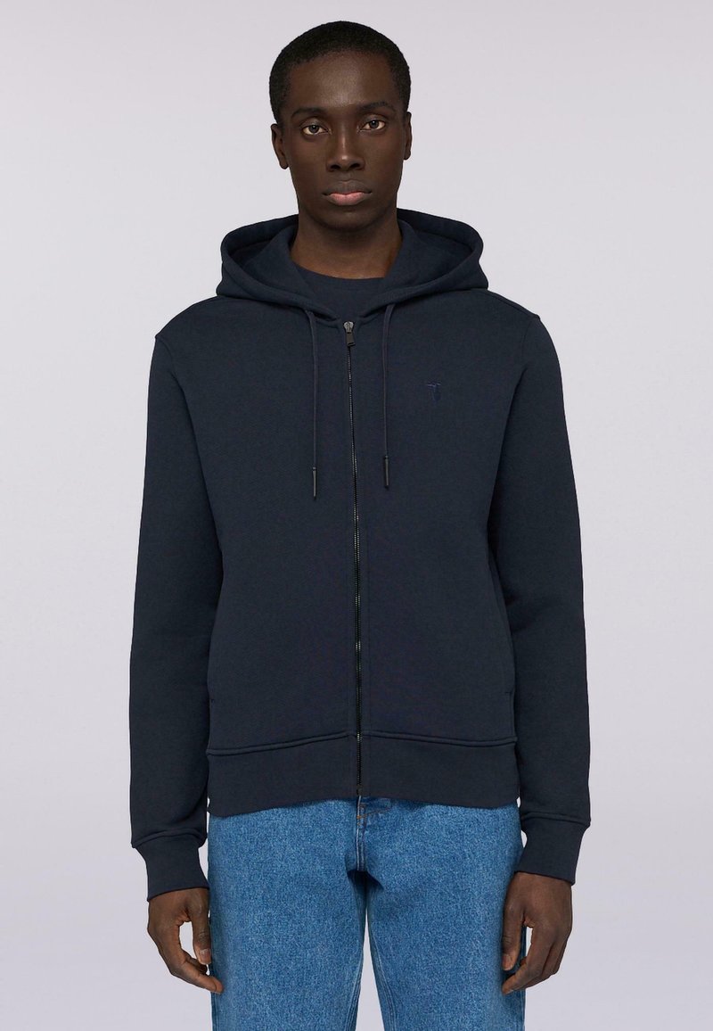 Námořnická mikina na zip z texturované tkaniny, s kapucí na stažení, dlouhými rukávy a decentním logem na hrudi. V kombinaci s modrými džíny.