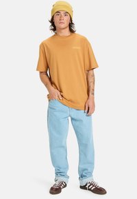 Sennepsgul kortærmet t-shirt med logo, parret med lyseblå løsthængende jeans og brune sneakers med hvide striber.