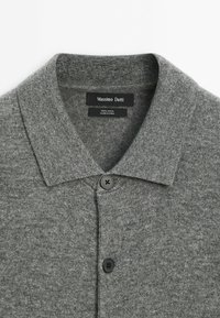 Cardigan en laine grise avec col et boutons noirs, étiquette Massimo Dutti, fabriqué en Chine, posé à plat sur une surface blanche.