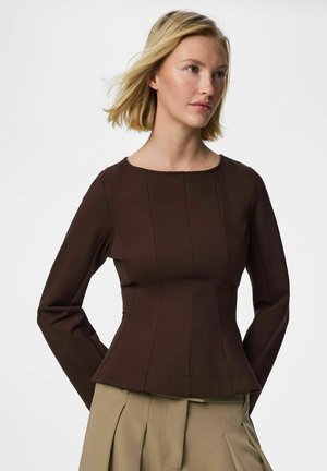 Blond kvinne med skulderlangt hår, iført en tettsittende mørkebrun langermet topp og beige plissert bukse, står foran en hvit bakgrunn.