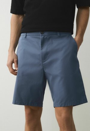 Man wearing mid-length blue shorts and a black tucked-in shirt, standing with one hand in pocket against a plain background.