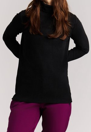 Personne aux longs cheveux bruns portant un pull à col roulé côtelé noir et un pantalon violet, debout, les mains derrière le dos.