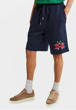 Man draagt marineblauwe korte broek met rood polo spelerlogo, witte sokken en bruine sneakers, staand met handen in zakken.