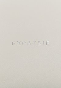 White leather surface featuring debossed silver text "EXPATRIÉ" with a textured finish. The material appears smooth but slightly grainy.
