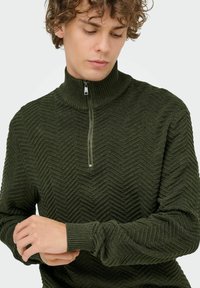 Dunkelgrüner Pullover mit hohem Kragen, mit einem strukturierten Fischgrätenmuster und einem halben Reißverschluss vorne. Weiches Strickmaterial.