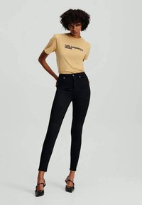 Valkoinen T-paita, jossa musta teksti "KARL LAGERFELD JEANS", yhdistettynä mustiin skinny-farkkuihin ja mustiin korkokenkiin, korostaen istuvaa siluettia.