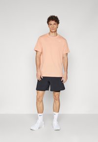 Conjunto deportivo que incluye una camiseta de manga corta de color durazno claro y pantalones cortos negros. Presenta un logo blanco de Nike en la camiseta. Zapatillas y calcetines blancos.