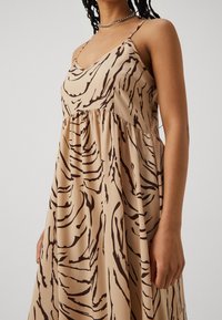 Vestido beige con una silueta A-line fluida. Presenta rayas oscuras similares a las de una cebra, tirantes finos ajustables y una cintura fruncida.