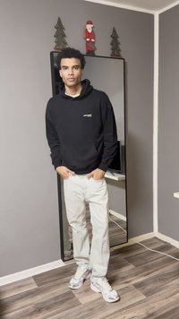 Sweat-shirt à capuche noir avec texte blanc, pantalon gris clair et baskets blanches. Debout près d'un miroir avec un mur gris et un sol en bois.