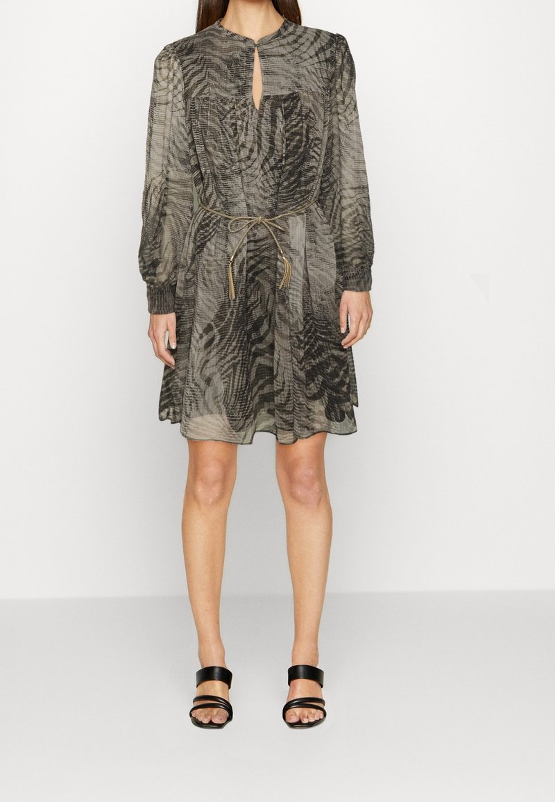 Mujer vestida con un vestido transparente de manga larga hasta la rodilla con un patrón abstracto oscuro, cinturón que ajusta la cintura y sandalias negras con tacón y dedos descubiertos.