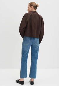 Braune, kurz geschnittene Jacke aus weichem Material, kombiniert mit hellblauen, geraden Jeans. Schwarze Slipper vervollständigen das Outfit.