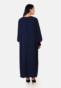 Robe longue bleu marine avec un décolleté rond, des manches longues avec des poignets froncés et une fermeture éclair au dos. Tissu lisse, design jusqu'au sol, style minimaliste.