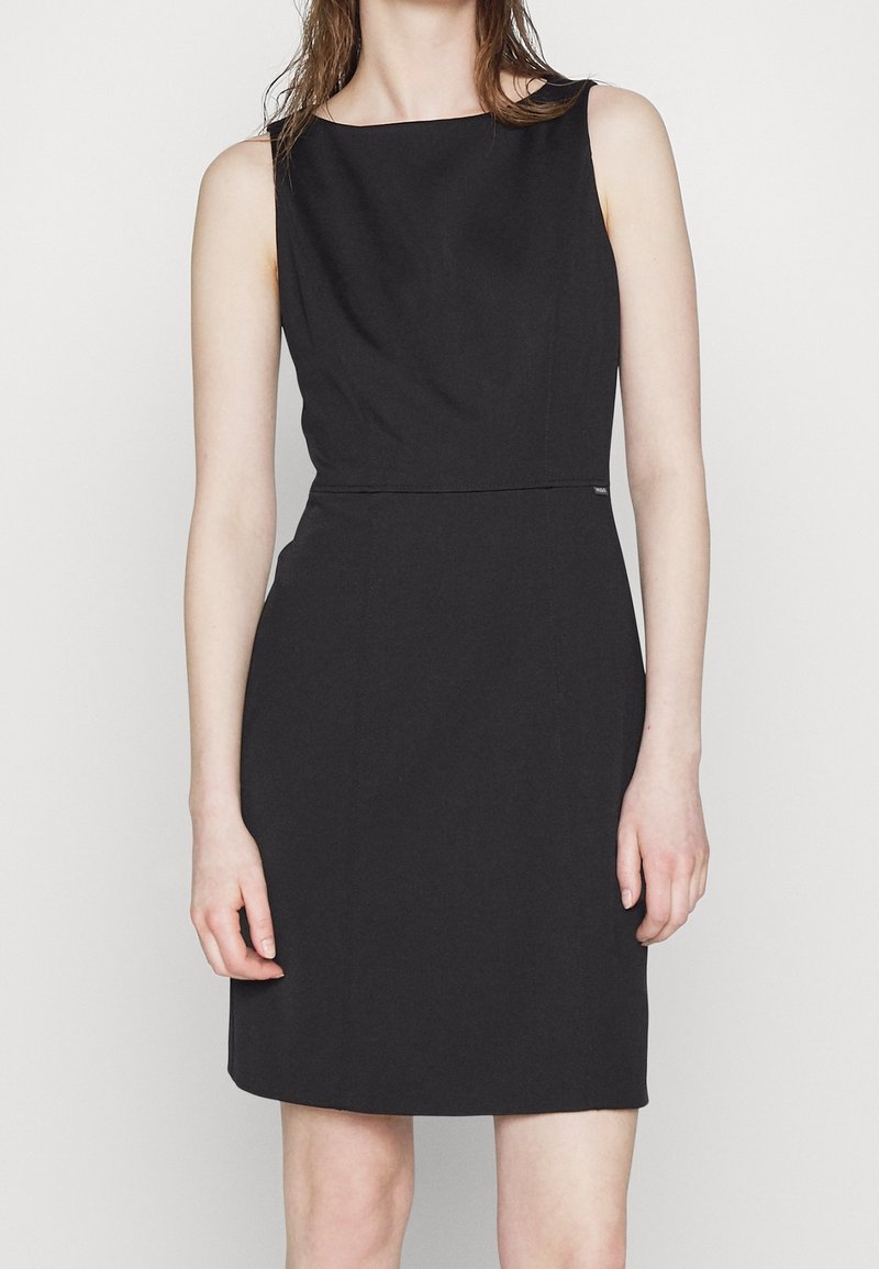 Robe noire sans manches longueur genou avec un col haut portée par une personne à la peau claire et aux cheveux bruns sur un fond uni.