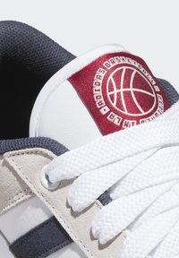 White sneaker featuring a red and white basketball logo on the tongue, white laces, and a mix of gray and beige materials with textured accents.